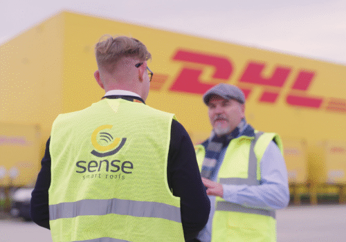 Dwóch pracowników w kamizelkach odblaskowych rozmawia na terenie DHL; na pierwszym planie widoczne logo SENSE Smart Roofs na plecach jednego z nich, w tle żółty budynek DHL.