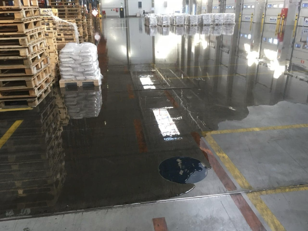 Zalana wodą po opadach deszczu posadzka wewnątrz hali , z paletami i składowanymi towarami.